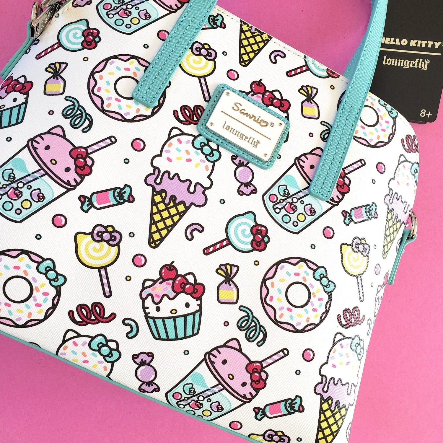 A multicolored Loungefly Hello Kitty Sweet Treats Crossbody Bag with character-themed candy and dessert patterns, displayed against a pink background.
