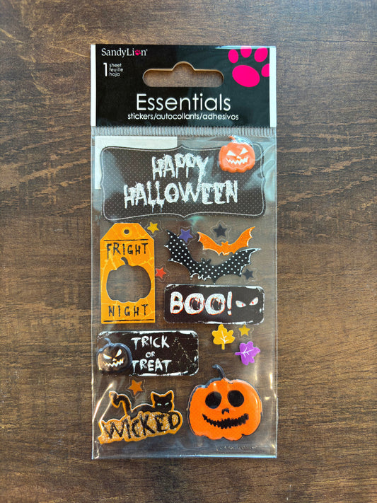 Sandy Lion Essentials Halloween sticker sheet in clear plastic packaging, featuring various spooky designs like pumpkins, bats, ghosts, and text stickers, displayed on a dark wood background.
