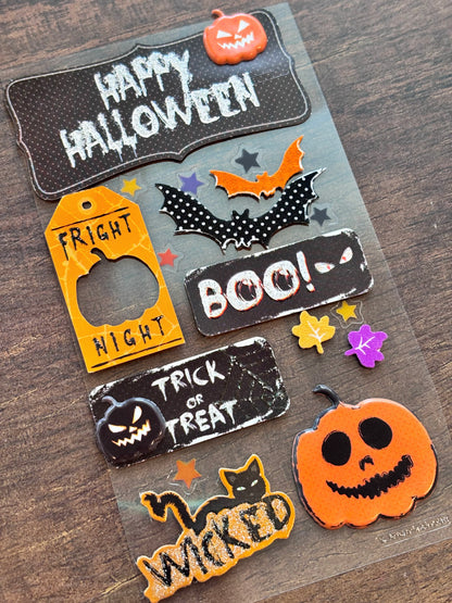 A sheet of Halloween-themed decorative stickers or decals arranged on a wooden surface. The designs include a large 'Happy Halloween' sign with a pumpkin, orange 'Fright Night' tags, black bats, 'BOO!' and 'Trick or Treat' signs, a black cat on 'Wicked' in glittery orange lettering, and a jack-o'-lantern face.