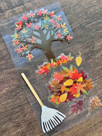 An autumn-themed craft set featuring a die-cut tree with colorful fall leaves, a pile of scattered autumn leaves, and a white rake with a wooden handle, all arranged on a clear sheet over a dark wood background.