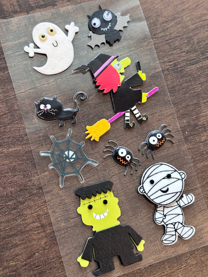 A sheet of Halloween-themed felt cutouts arranged on a dark wood surface, including a ghost, black cat, witch, spider, mummy, Frankenstein's monster, spiderweb, and black bat.
