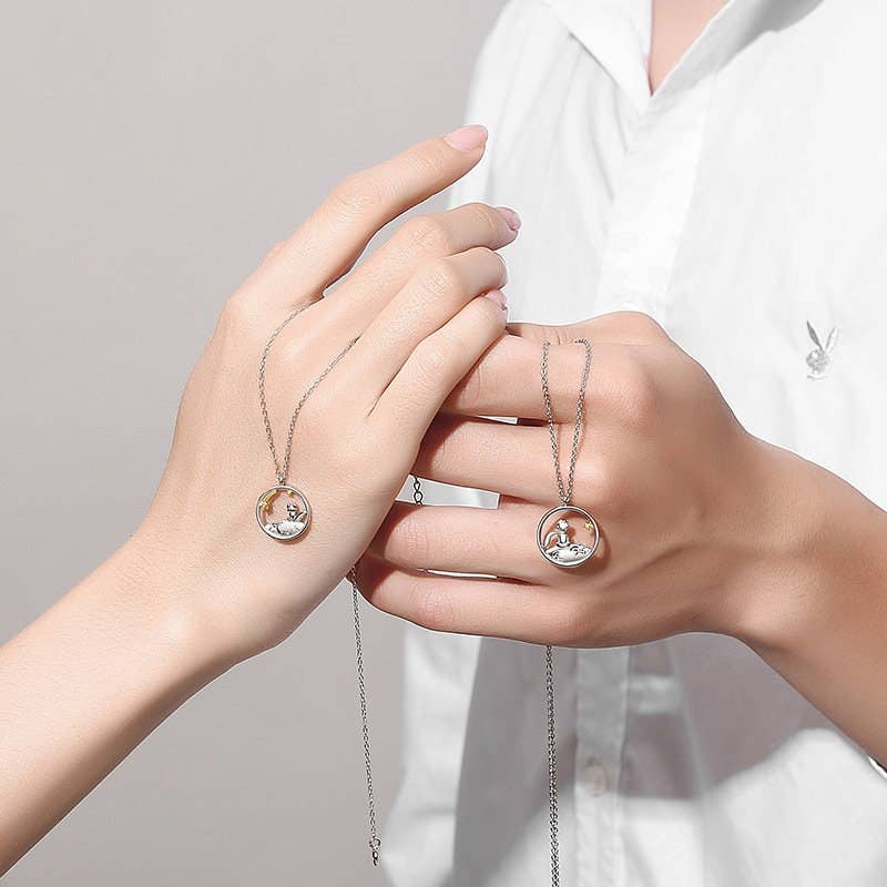 Two hands holding matching The Little Prince silver necklaces with circular pendants against a neutral background