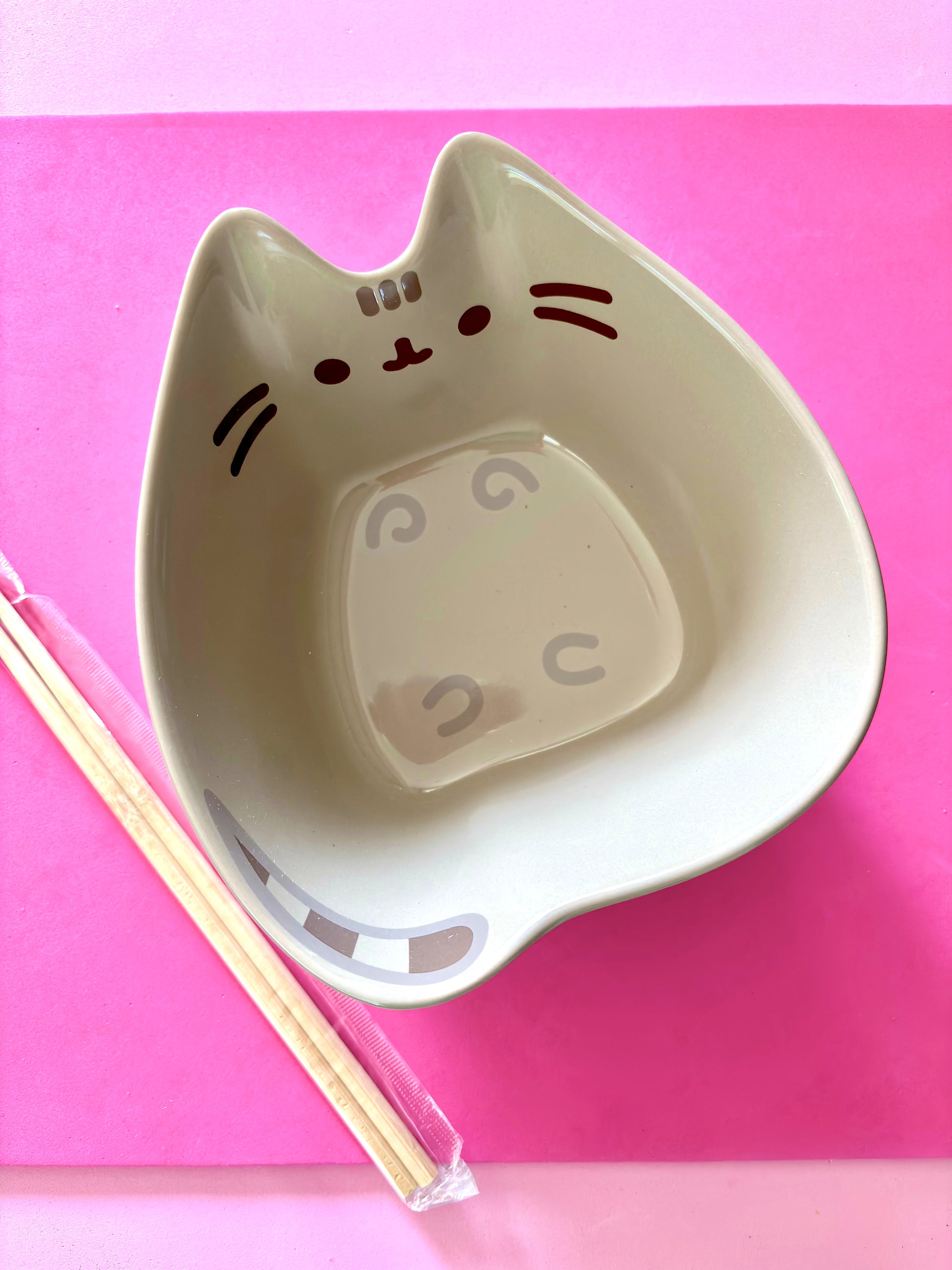 A cat-shaped ceramic bowl in a gray color with a cute face printed on the inside, shown on a bright pink background with a pair of chopsticks resting beside it.