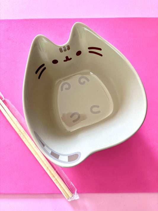 A cat-shaped ceramic bowl in a gray color with a cute face printed on the inside, shown on a bright pink background with a pair of chopsticks resting beside it.