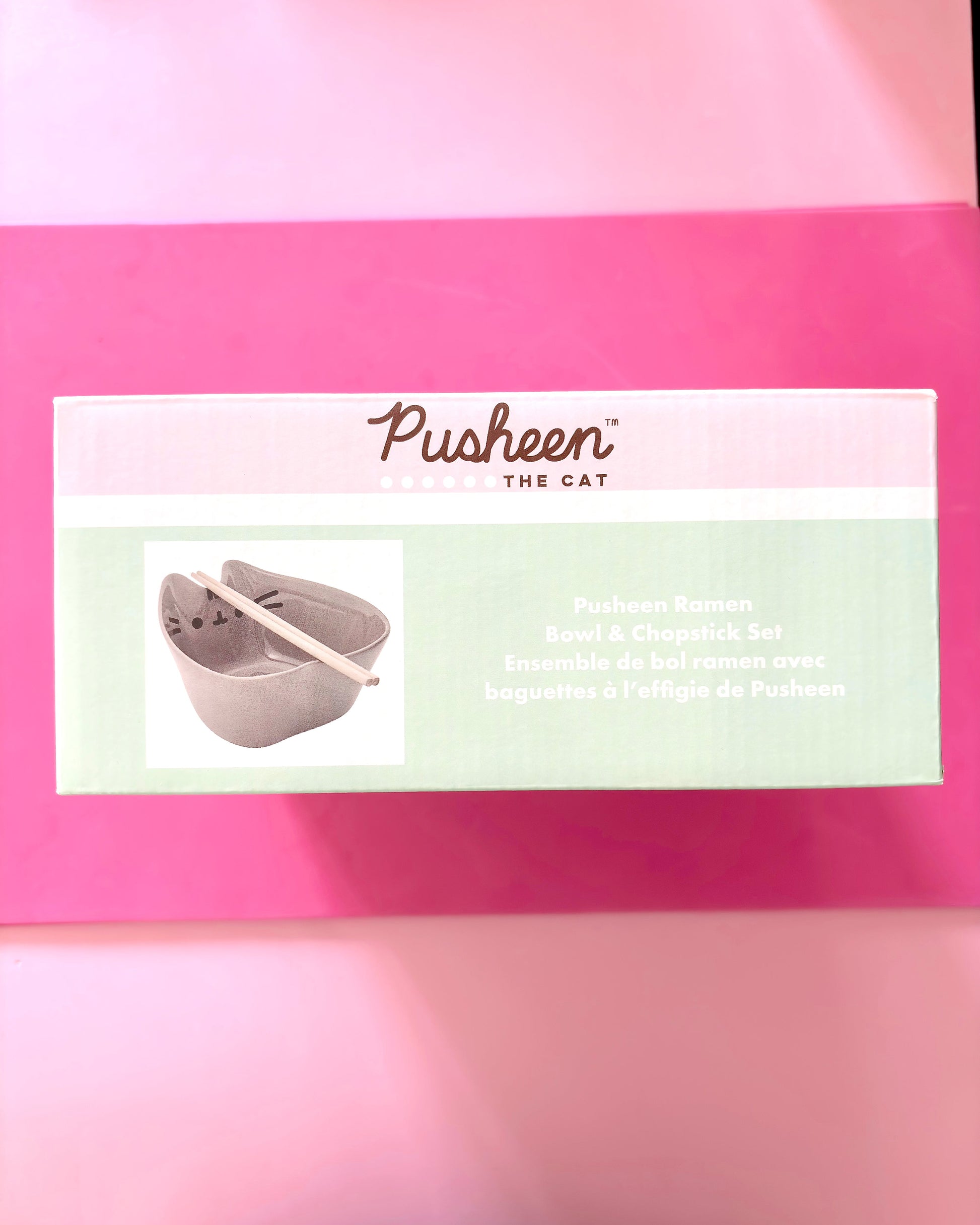 A boxed Pusheen The Cat Pushkin Ramen Bowl & Chopstick Set placed on a bright pink background. The front of the package shows the Pusheen branding and a small photo of the gray cat-shaped bowl with chopsticks.