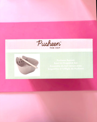 A boxed Pusheen The Cat Pushkin Ramen Bowl & Chopstick Set placed on a bright pink background. The front of the package shows the Pusheen branding and a small photo of the gray cat-shaped bowl with chopsticks.